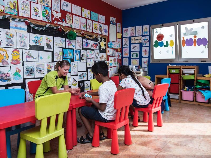 Tre bambini seduti a tavoli colorati in una classe con disegni su pareti blu scuro e rosse.