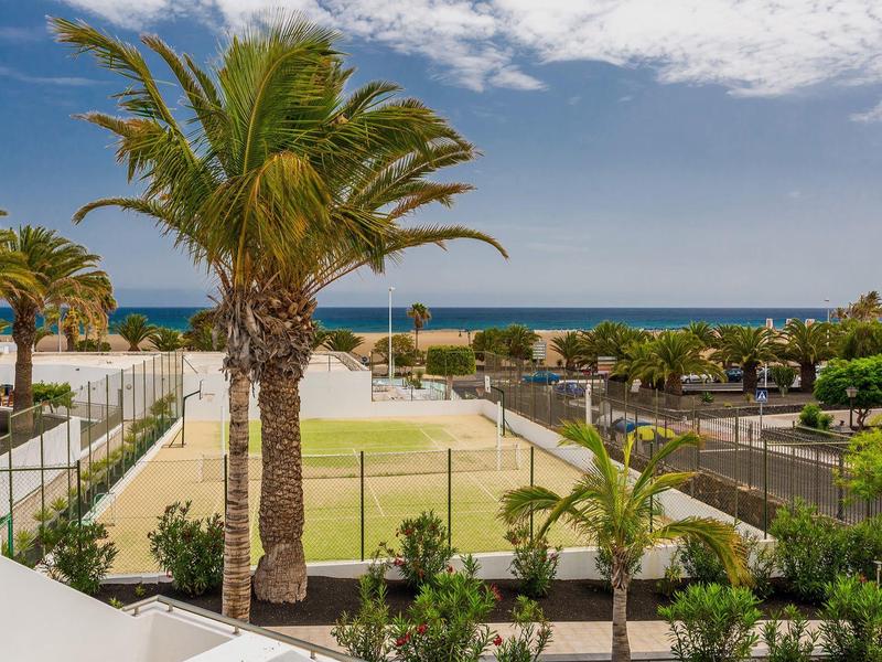 Vista costiera con palme, campi sportivi verdi e oceano blu sotto un cielo parzialmente nuvoloso.
