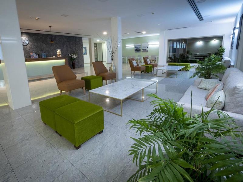 Lounge d'hotel moderna con divani bianchi, pouf verdi, tavoli in vetro, piante e pavimento piastrellato.