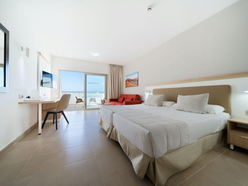 Camera d'hotel luminosa con due letti, scrivania, sedia e balcone con vista mare.