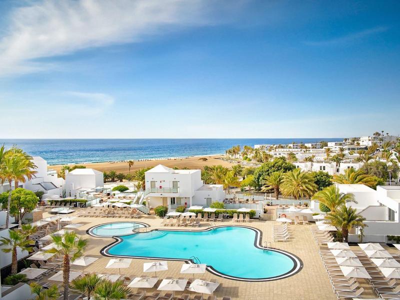 Resort con grande piscina circondata da lettini bianchi e ville bianche vicino all'oceano.