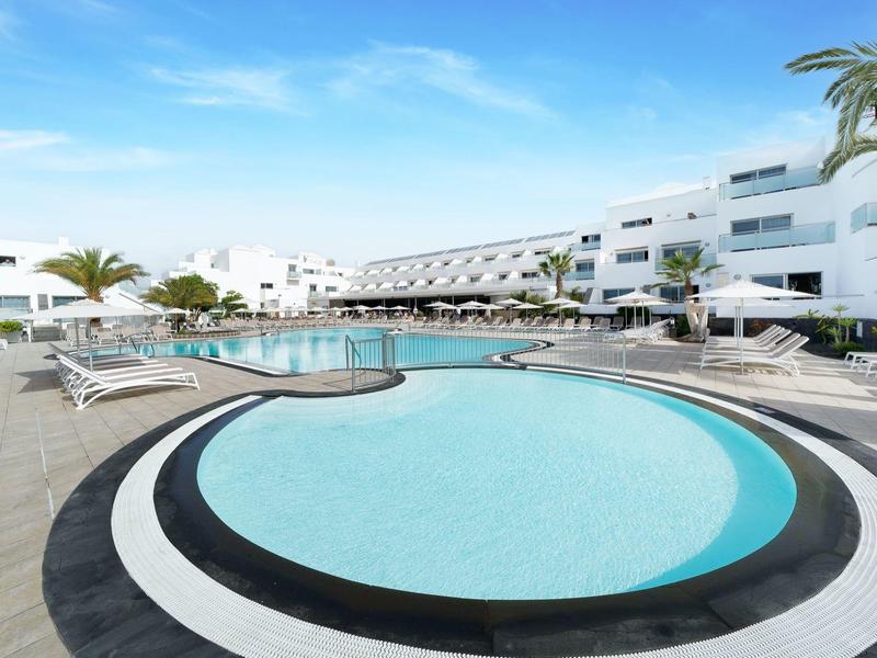 Area piscina esterna dell'hotel con palme, lettini e edifici moderni bianchi.