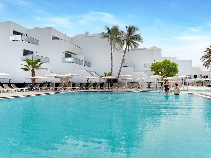 Hotel bianco moderno con piscina, lettini e palme sotto un cielo azzurro.