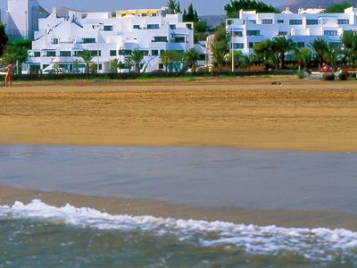 Weißes Hotel in Strandnähe mit Palmen, Sand und sanften Wellen im Vordergrund.