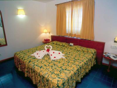 Ein Schlafzimmer mit Doppelbett, gelb-grün gemustert, Ornamenten, rotem Kopfteil und Vorhängen.