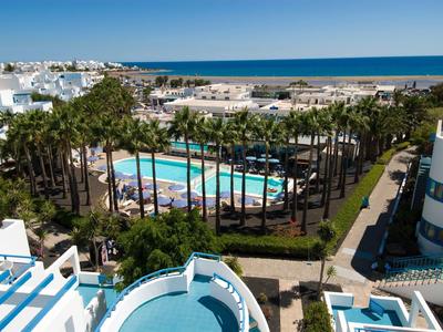 Blick auf Hotel mit Pool, Palmen, weißen Gebäuden und dahinter das blaue Meer unter klarem Himmel.