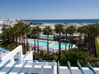 Blick auf einen Pool mit Palmen, dahinter Sandstrand und blaues Meer unter klarem Himmel.