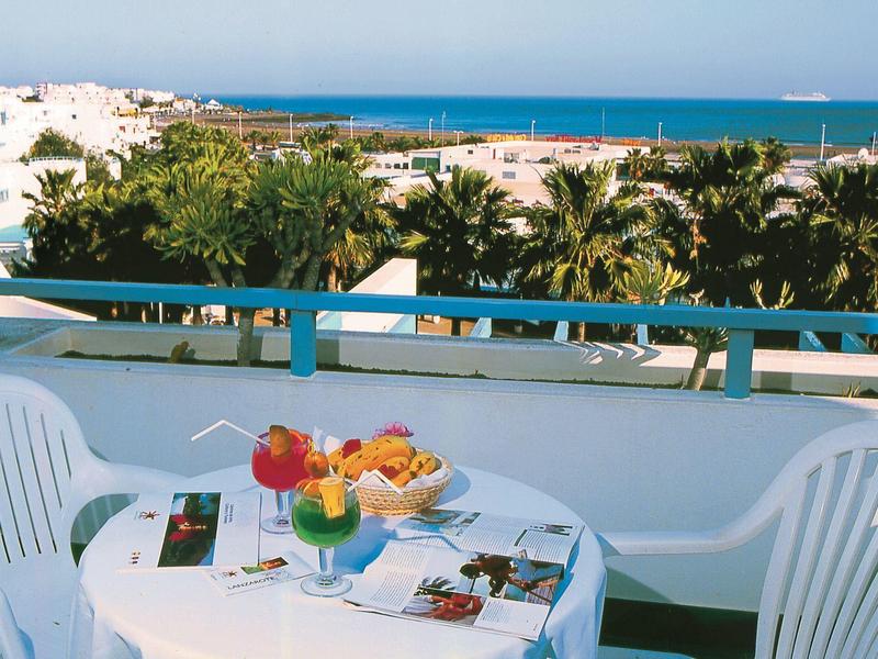 Balcone con tavolo, sedie, cibo e vista su palme e mare.