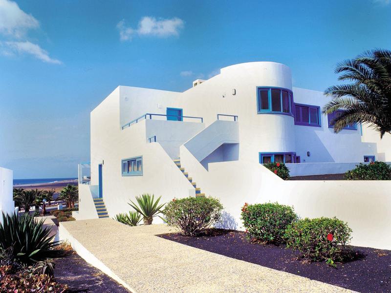 Edificio bianco moderno con terrazza e piante in ambiente costiero soleggiato.