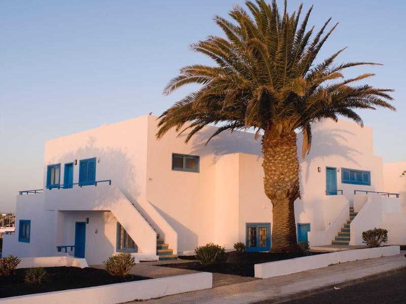 Casa vacanze bianca con finestre e porte blu, circondata da una palma e cielo azzurro.