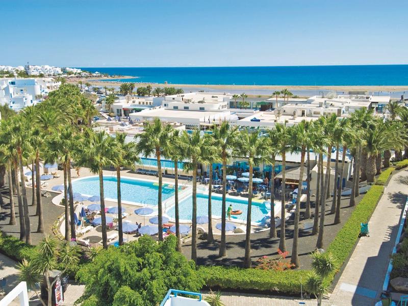 Hotel con piscina circondata da palme, vista sul mare azzurro sullo sfondo.