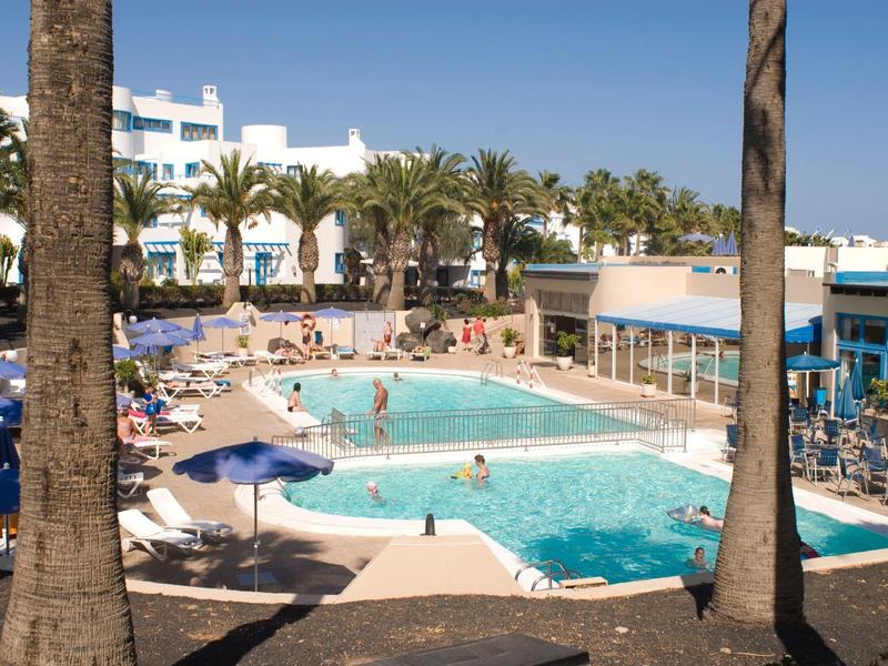 Piscina dell'hotel con ospiti, ombrelloni e palme sotto un cielo limpido.