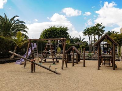 Parco giochi per bambini con strutture per arrampicata e sabbia sotto palme con cielo azzurro.
