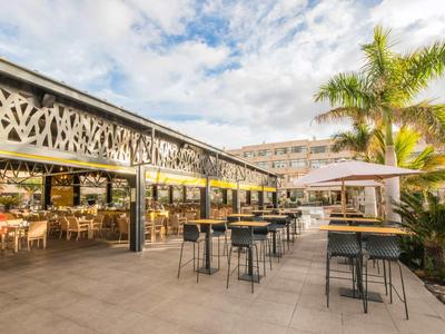 Moderna terrazza dell'hotel con tavoli, sedie e palme sotto un cielo nuvoloso.