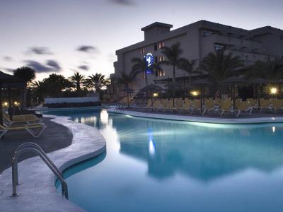 Piscina dell'hotel illuminata al crepuscolo con lettini e palme sullo sfondo.