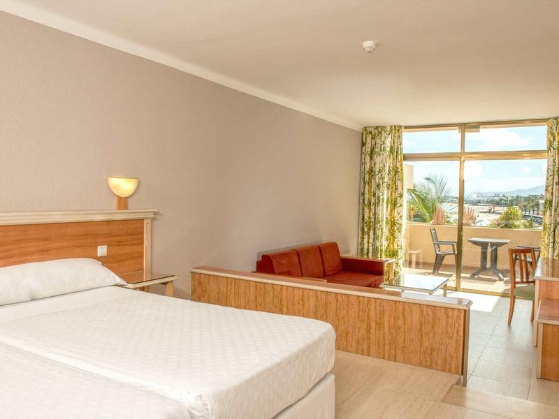 Camera d'hotel luminosa con letto matrimoniale, divano e balcone con vista su un paesaggio verde.