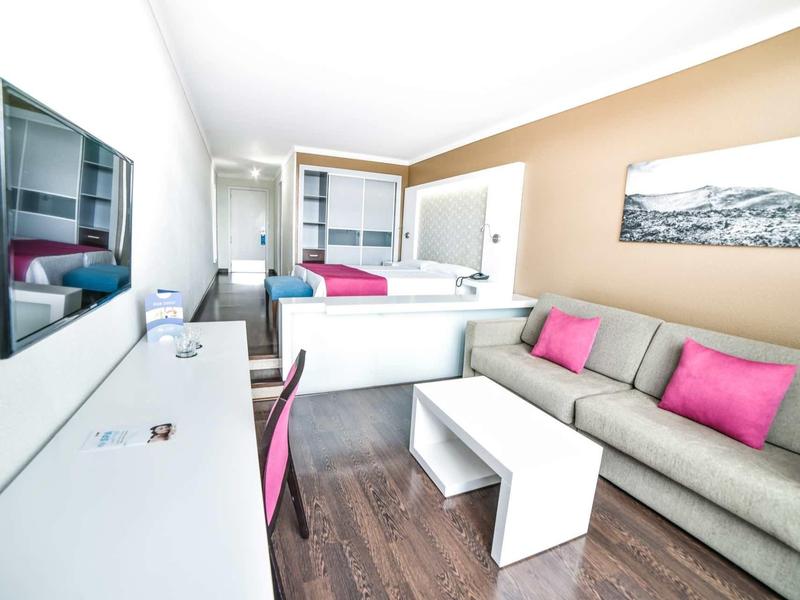 Camera d'hotel moderna con divano, TV e area lavoro in colori chiari.