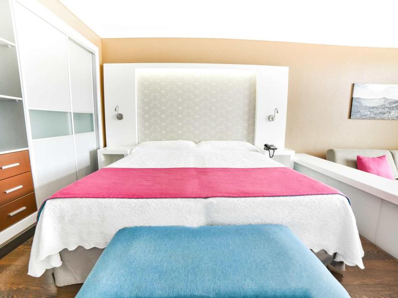 Camera d'hotel luminosa con letto grande, coperta rosa e panca blu ai piedi del letto.