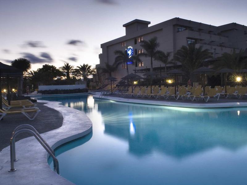 Piscina dell'hotel illuminata al crepuscolo con lettini e palme sullo sfondo.