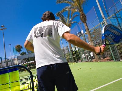 Mann spielt Padel auf grünem Platz mit Palmen und blauem Himmel im Hintergrund.