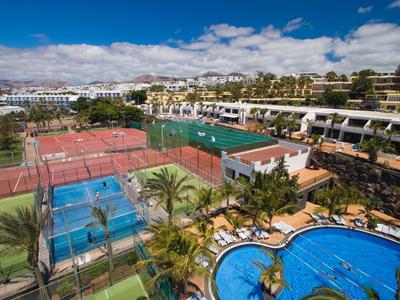 Freizeitgelände mit Tennisplätzen, Swimmingpool, Palmen und Gebäuden unter blauem Himmel.