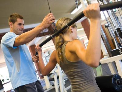 Frau zieht Lat-Zugstange, Trainer unterstützt dabei im Fitnessstudio.