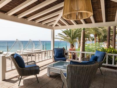 Gemütliche Terrasse mit blauen Sesseln, Holztisch, überdacht, Blick auf Meer und Palmen.