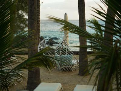 Zwei weiße Liegestühle mit kleinem Tisch stehen unter Palmen am Strand, im Hintergrund das Meer.