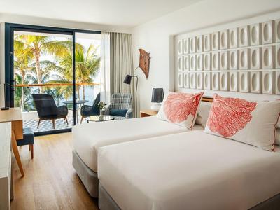 Hell eingerichtetes Hotelzimmer mit zwei Betten, Balkon und Blick auf Palmen und Meer.