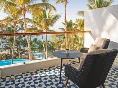 Balkon mit blauem Fliesenboden, Korbstuhl, kleinem Tisch, Palmen und Pool im Hintergrund.
