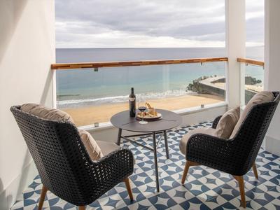 Balkon mit zwei geflochtenen Stühlen, einem Tisch, Weinflasche und Meerblick bei bewölktem Himmel.