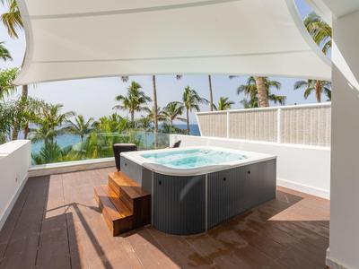 Moderner Whirlpool auf Holzterrasse unter Sonnensegel mit Palmen und Meer im Hintergrund.