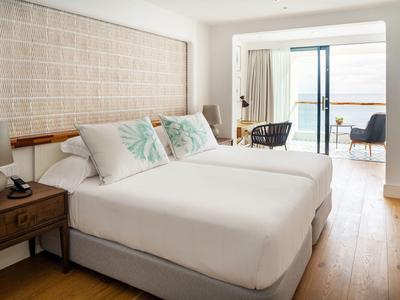 Helles Schlafzimmer mit zwei Betten, Holzboden, Balkon und Meerblick durch große Fenster.