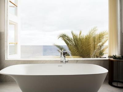 Weiße freistehende Badewanne vor offenem Fenster mit Meerblick und Palmen draußen