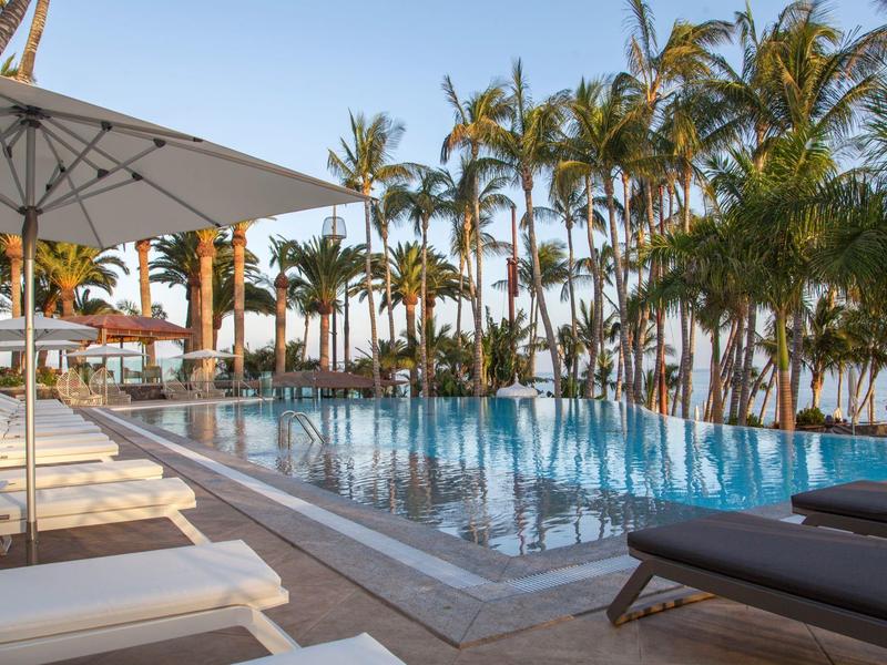 Una zona piscina tranquilla con lettini, ombrelloni e palme vicino al mare.