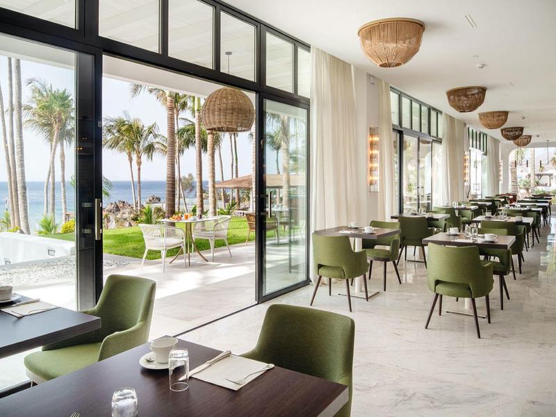 Ristorante luminoso con sedie verdi e grandi finestre che si affacciano su palme e mare.