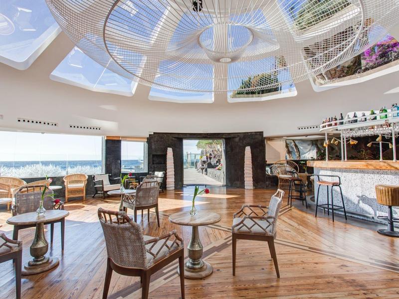 Bar dell'hotel luminoso con pavimento in legno, tavoli rotondi, sedie e grande tetto in vetro con vista.