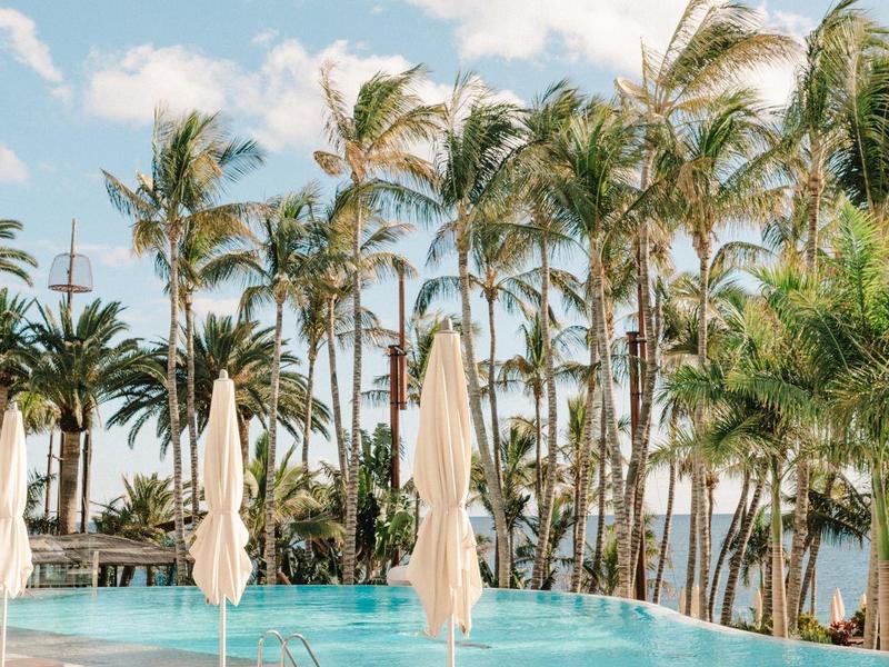 Lettini vuoti e ombrelloni chiusi accanto a una piscina con palme sullo sfondo.