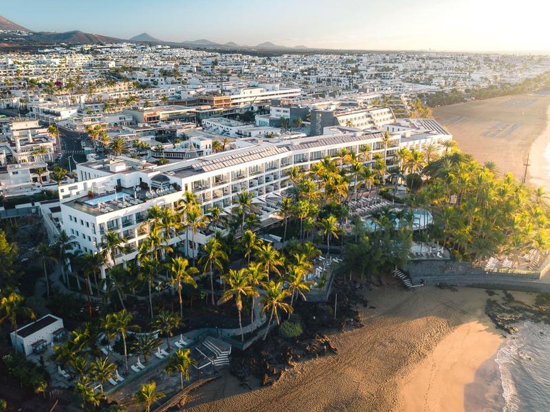 Grande hotel sulla costa con palme, spiaggia e città sullo sfondo al tramonto.