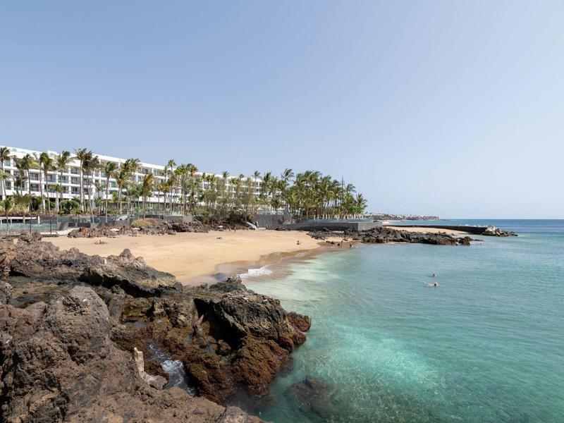 Costa con rocce, spiaggia sabbiosa e hotel sotto un cielo azzurro.