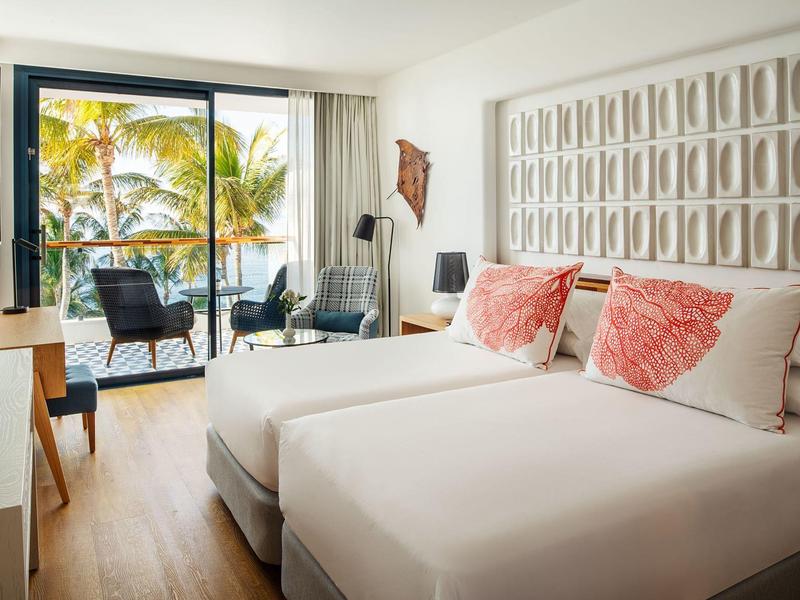 Camera d'hotel moderna con due letti e balcone con vista piscina e giardino.