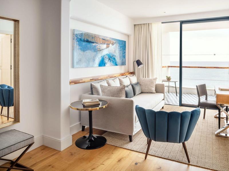 Camera d'hotel luminosa con poltrone blu, divano e ampia finestra con vista sul mare.