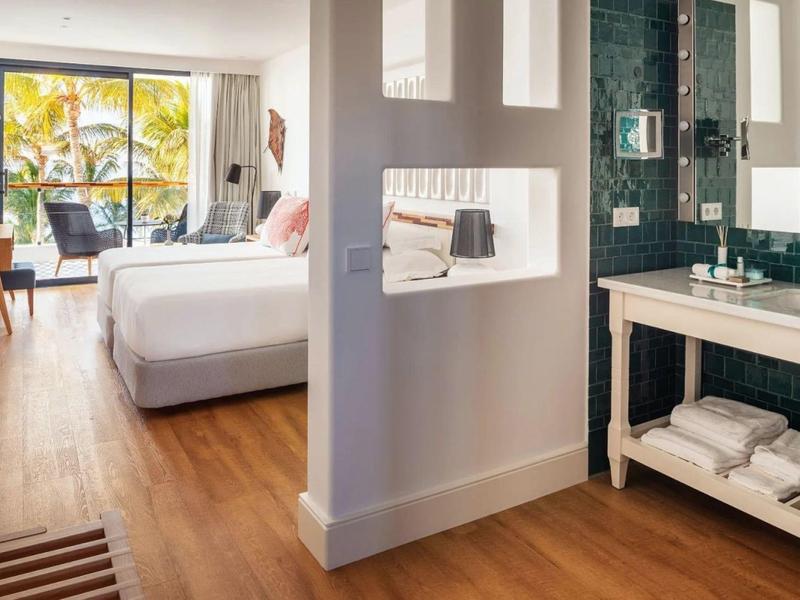 Camera d'hotel luminosa con letto matrimoniale, pavimento in legno e bagno adiacente con due lavandini.