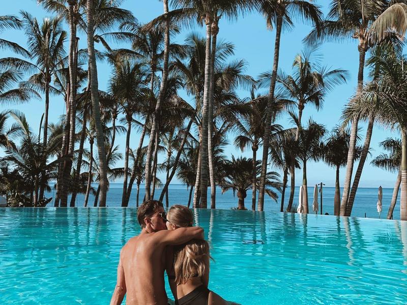 Coppia rilassata in piscina con palme e mare sullo sfondo in una giornata soleggiata.