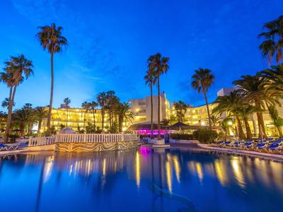 Großer beleuchteter Pool bei Nacht mit Palmen und Hotel im Hintergrund, klare blaue Wasserfläche.