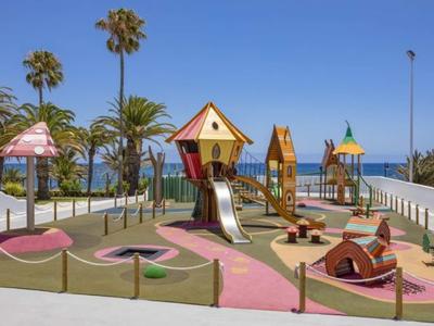 Bunter Spielplatz mit Rutschen und Klettergerüsten, umgeben von Palmen unter blauem Himmel.
