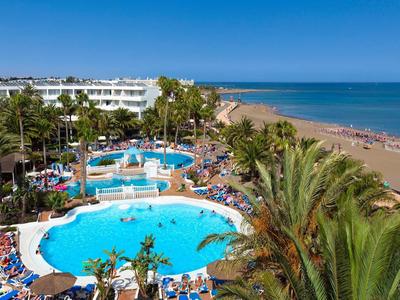 Hotelanlage mit großem Pool, grünen Palmen, Strand und blauem Meer im Hintergrund unter klarem Himmel.