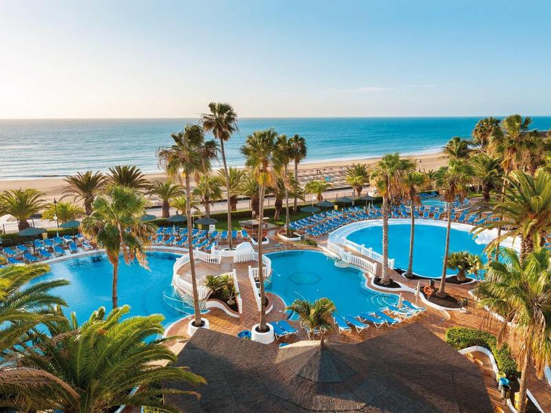 Piscina lussuosa circondata da palme con vista sulla spiaggia e sul mare al tramonto.