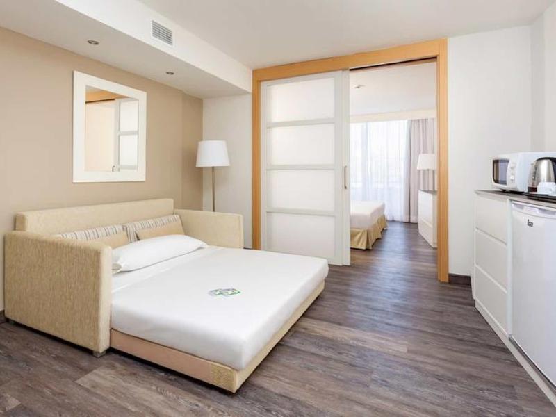 Monolocale d'hotel moderno con divano letto e angolo cottura, pavimento in legno scuro, luce naturale.