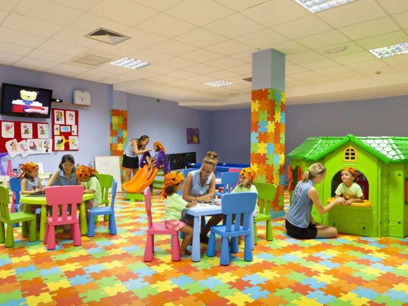 Area giochi colorata per bambini con tavoli, sedie e casetta verde.
